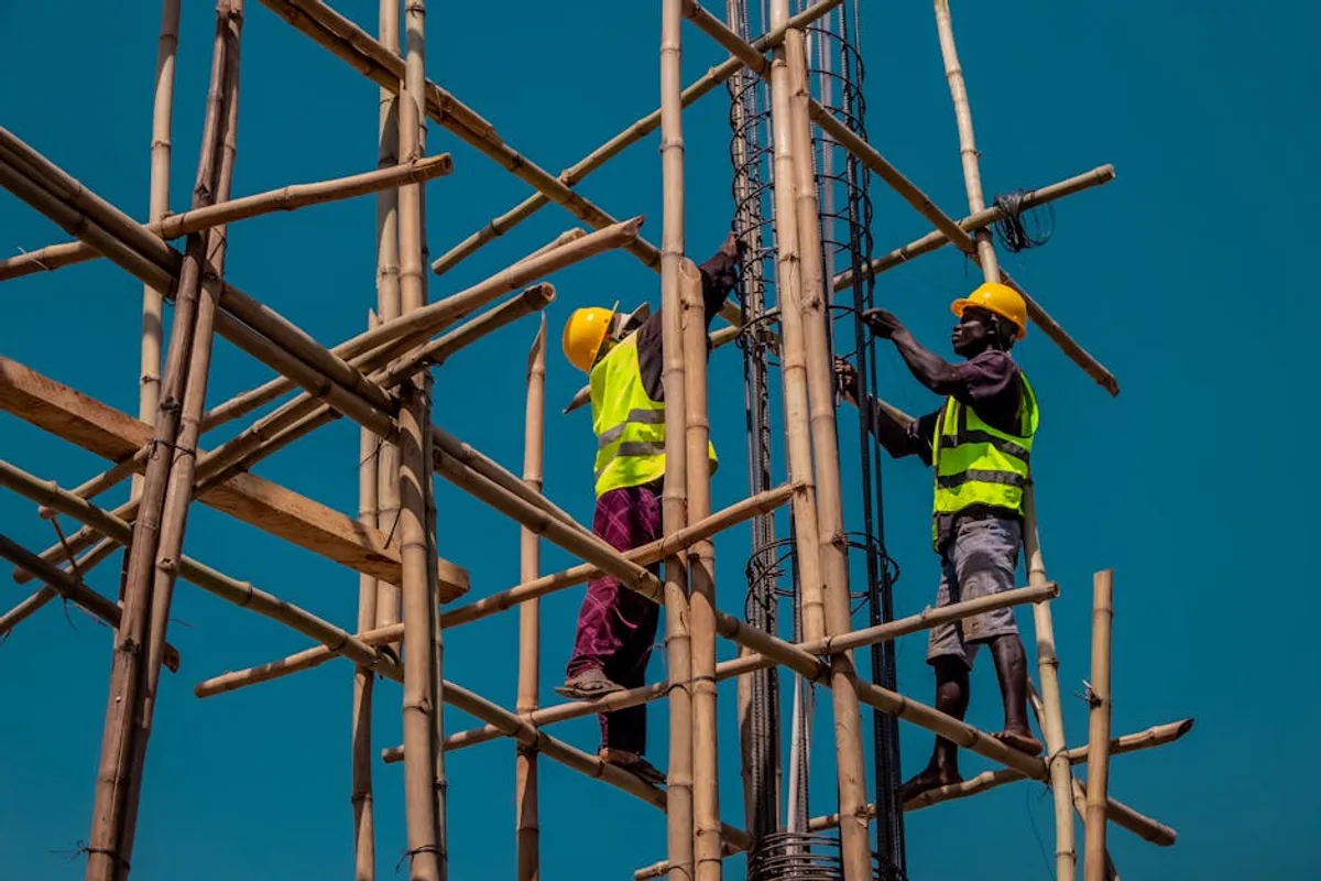 Two construction workers on bamboo scaffolding, wearing safety gear under a clear blue sky.