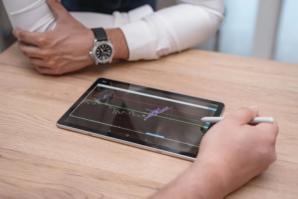 Close-up of hands using a digital tablet with trading charts on a wooden desk.