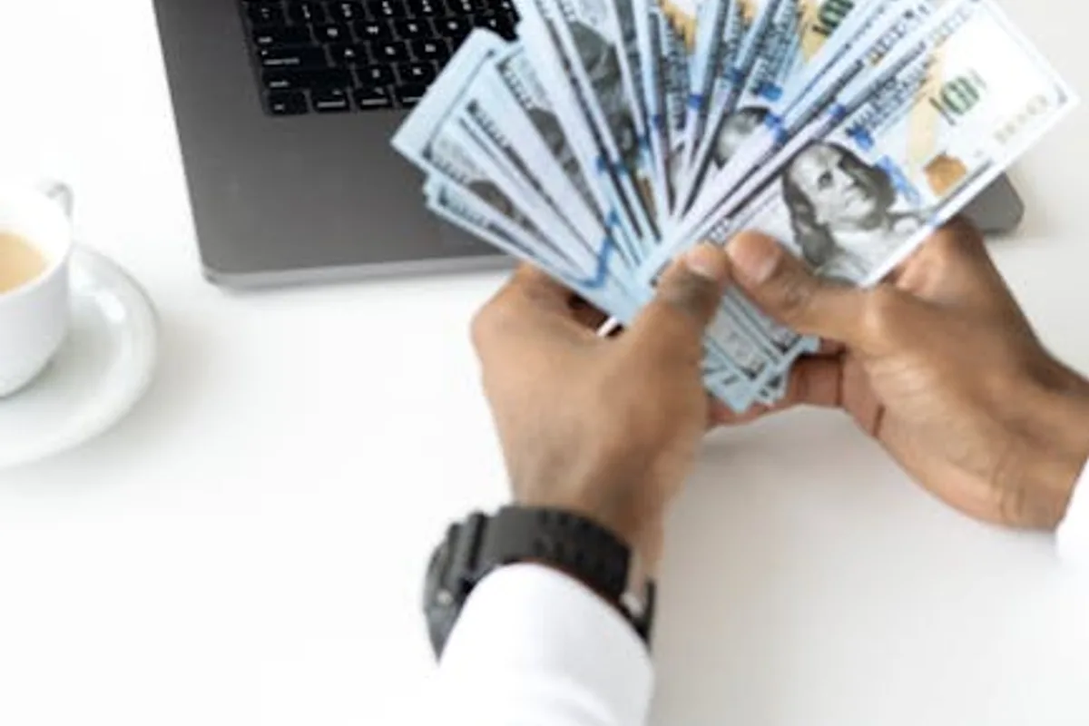 Hands holding cash in front of a laptop displaying stock market data, symbolizing financial growth and investment.