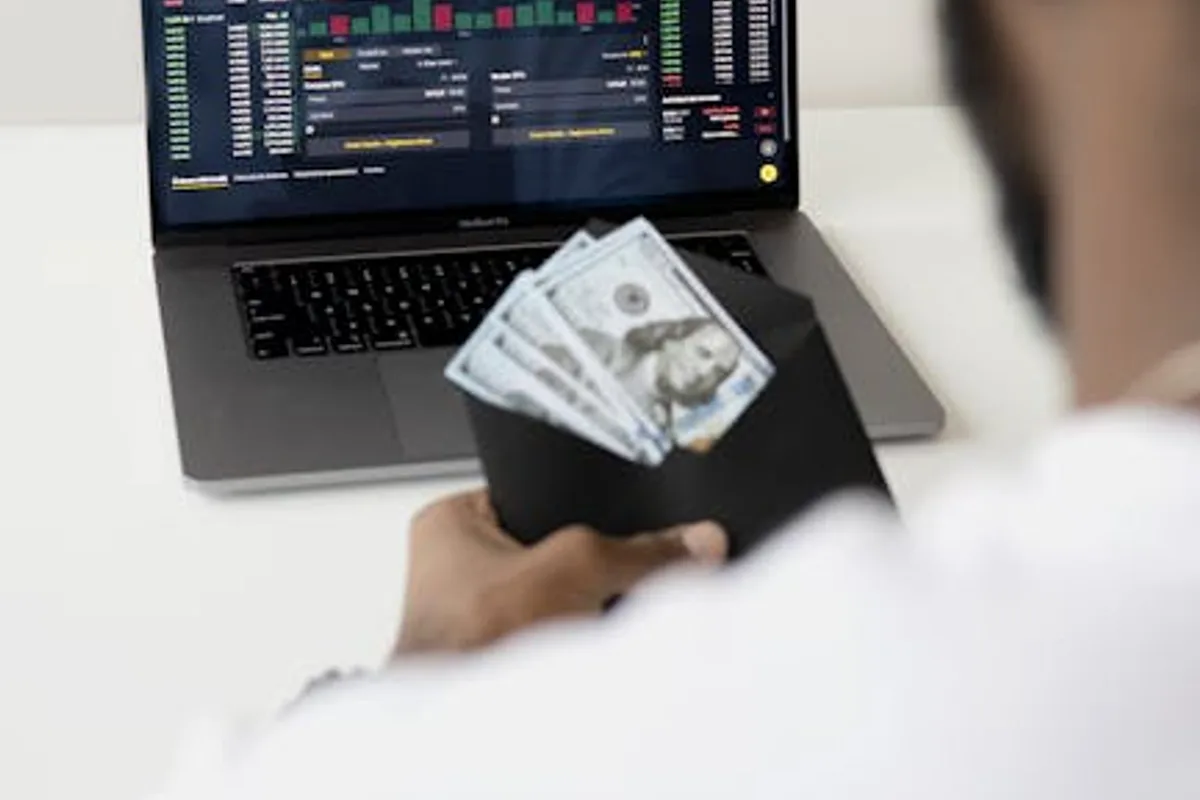 A person examines stock market data on a laptop while holding dollar bills, showcasing finance and trading concepts.